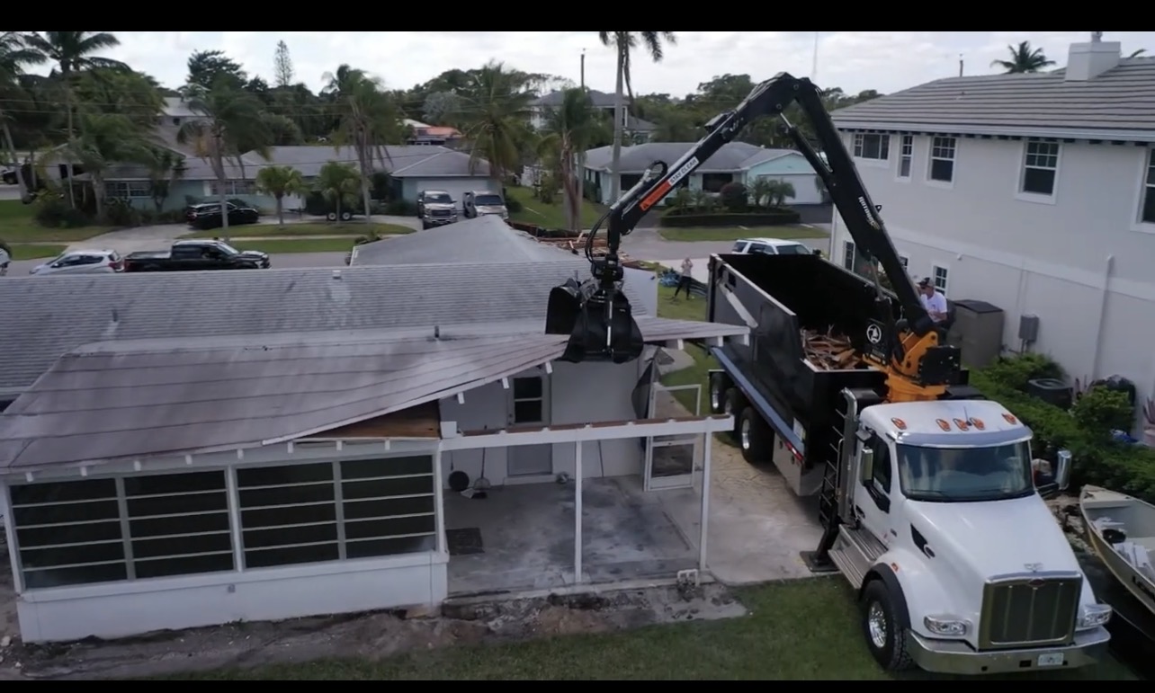 Grapple dump truck loading structure demolition debris beside a residential building
